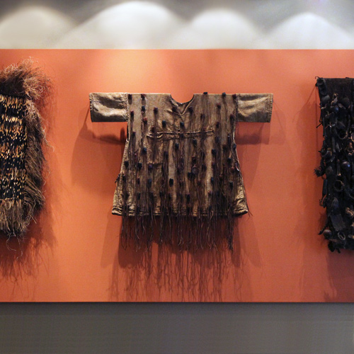 An African tunic hung against a terracotta-red wall, flanked by fringed fiber garments