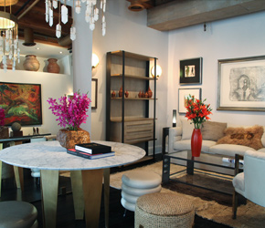 Light-filled room with a round marble-topped table, magenta orchids, open shelving of vessels and framed drawings