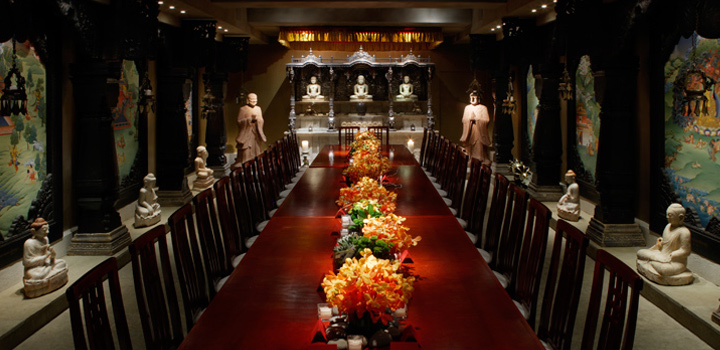 A long dining table set with orange flowers and dark wooden chairs in the Buddha Room, lined with Buddhas and painted panels
